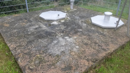 Verwitterte Betonfläche mit Bewuchs, zwei Metallabdeckungen und einer Rohrdurchführung.