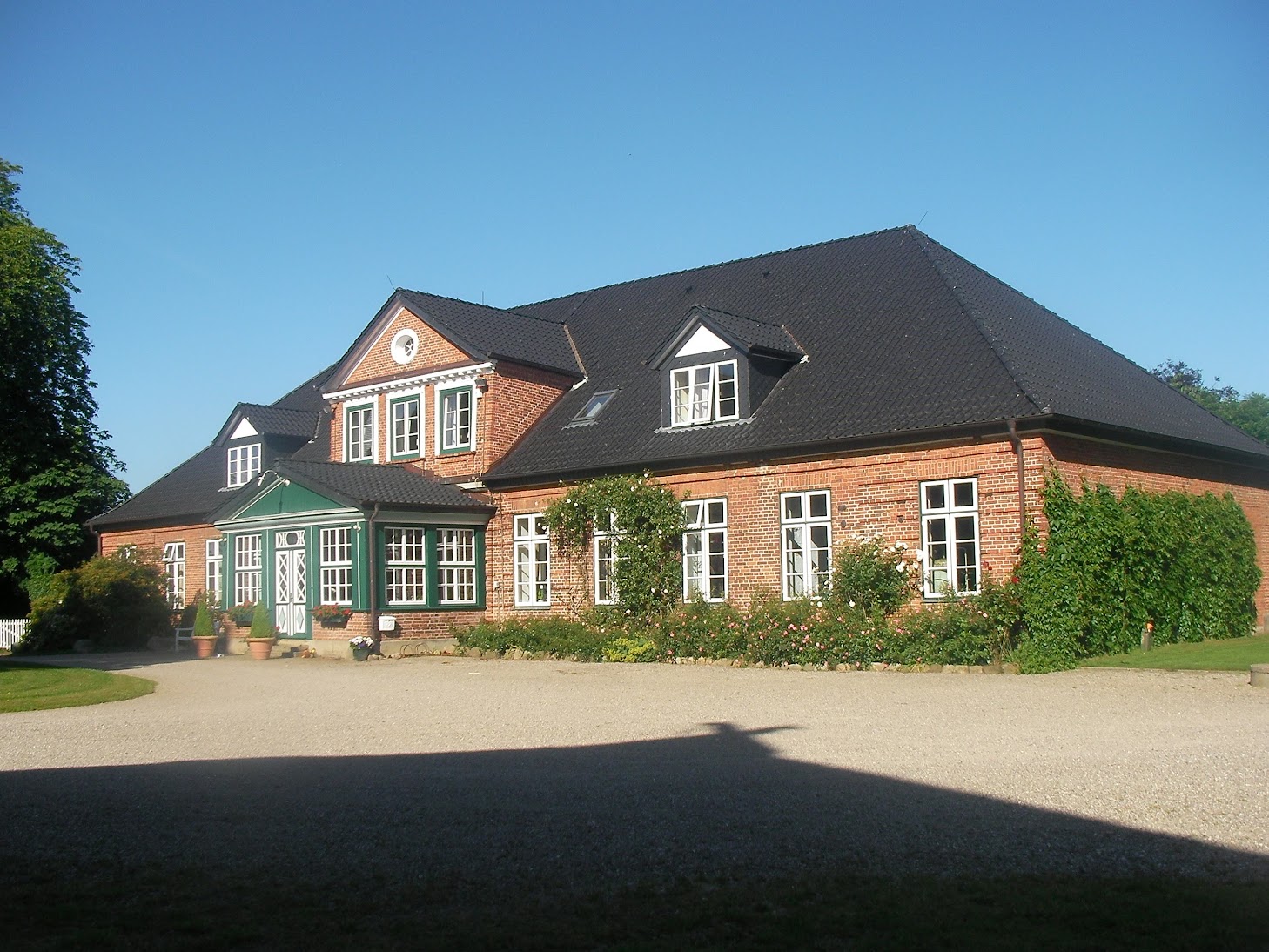 Großes Backsteinhaus mit dunklem Walmdach, Dachgauben und mittigem Zwerchgiebel.