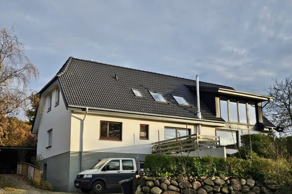 Weißes Wohnhaus mit dunkler Dacheindeckung, drei Dachfenstern und einer großen seitlichen Gaube.