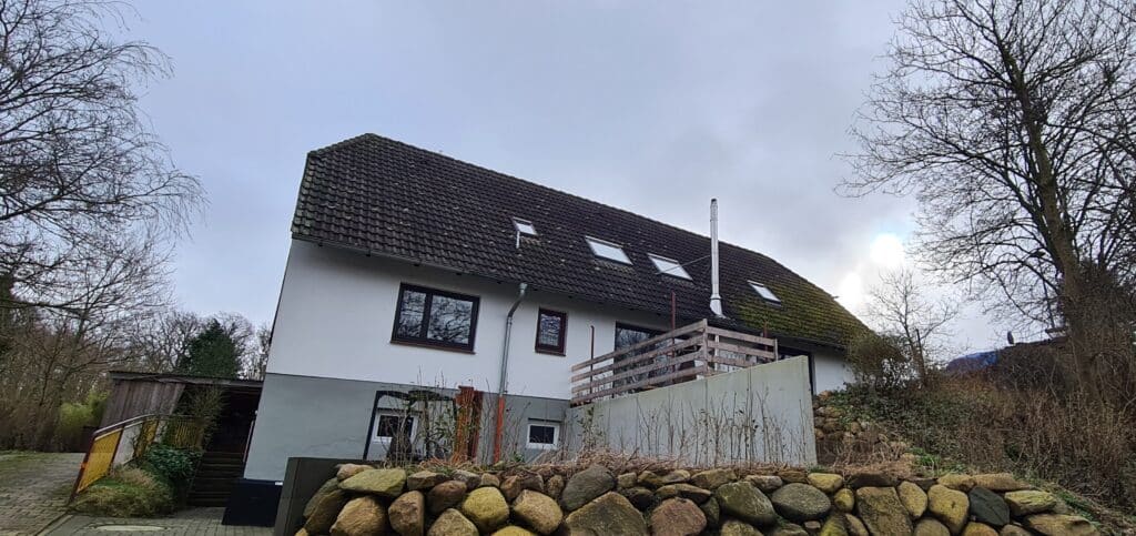 Weißes Wohnhaus mit älterem dunklen Dach, Dachfenstern und Terrasse vor der Dachsanierung.