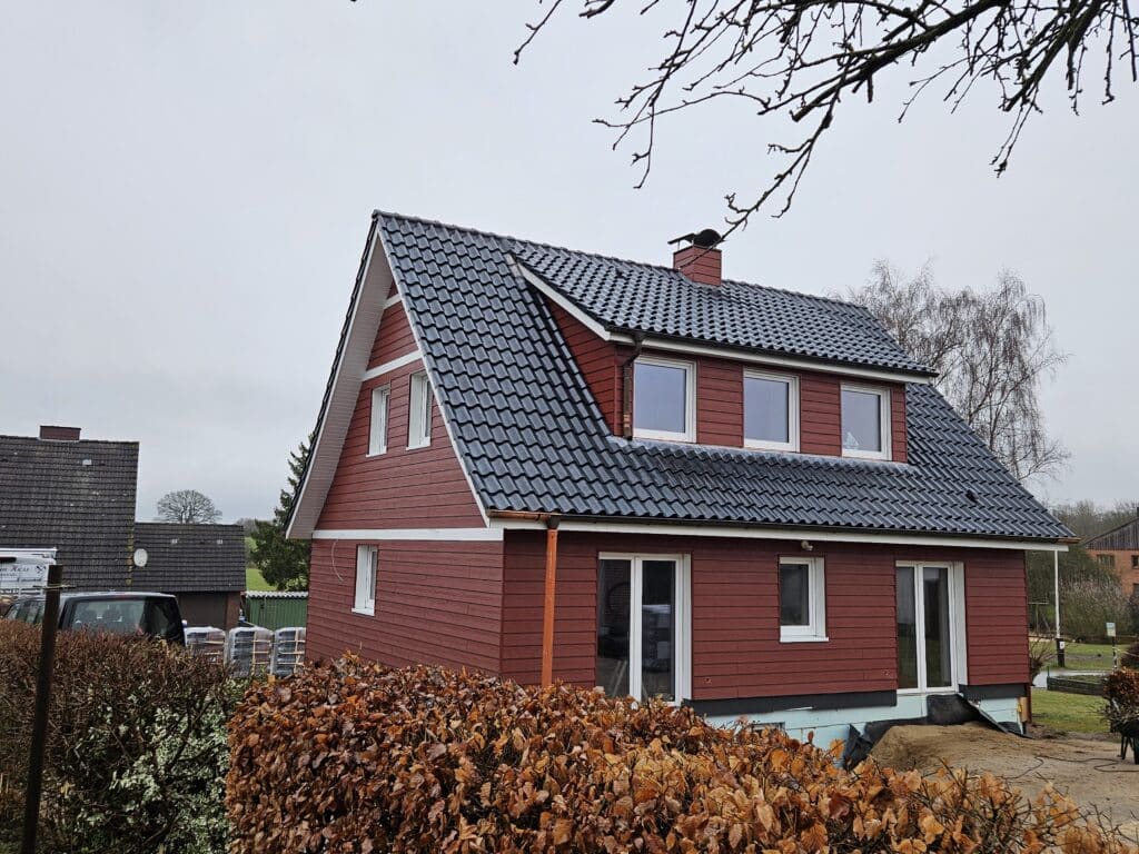 Wohnhaus mit roter waagerechter Fassadenverkleidung, weiß gerahmten Fenstern und dunklem Dach.
