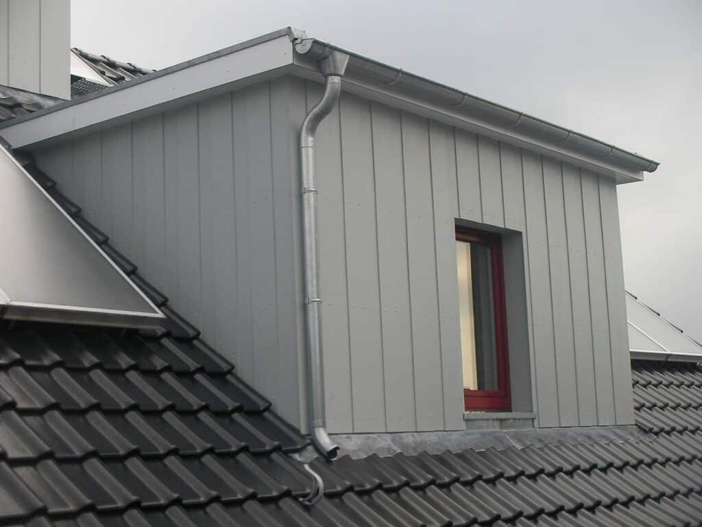 Dachgaube mit heller Metallverkleidung, Fenster, Dachrinne und Fallrohr.