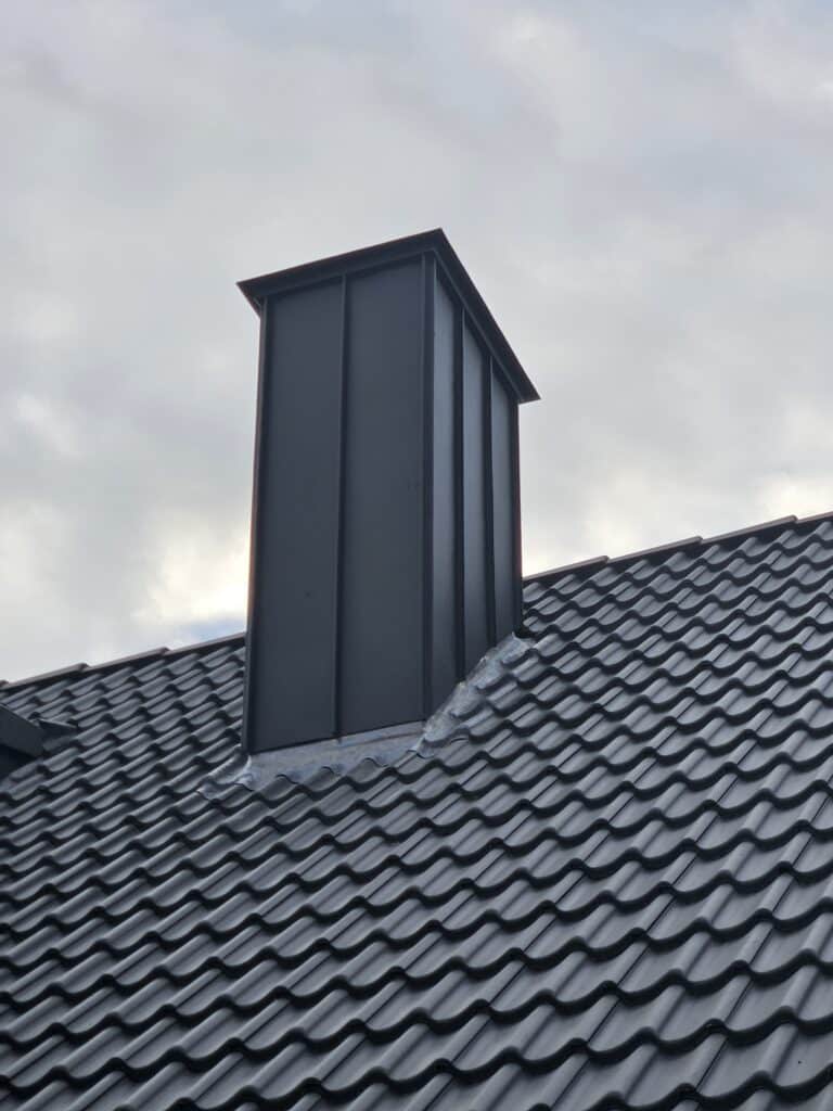 Schornstein mit dunkler Metallverkleidung und Abdeckung auf einem dunklen Ziegeldach.