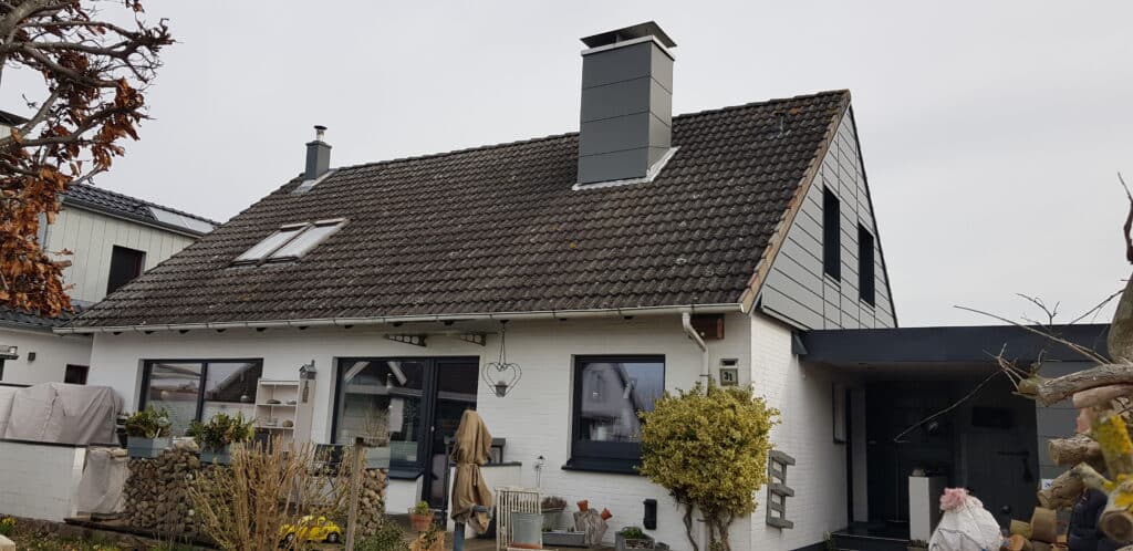Weißes Wohnhaus mit älterer Dacheindeckung und verkleideten Schornsteinen vor der Dachsanierung.
