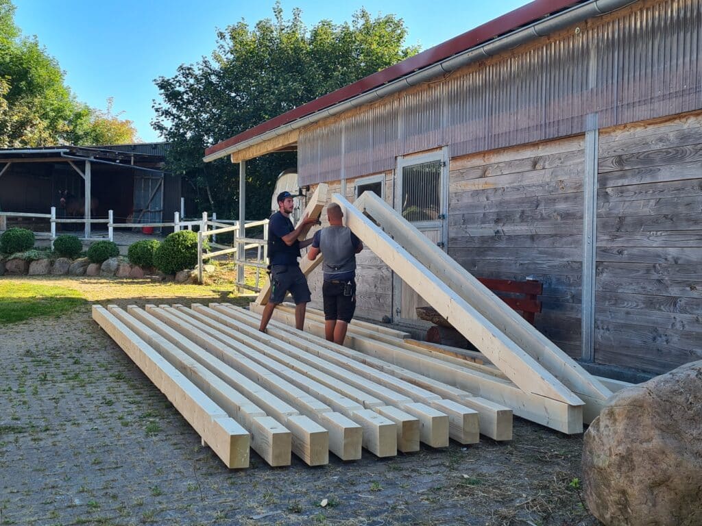Zwei Handwerker bearbeiten lange Holzbalken auf dem Betriebshof neben einem Holzgebäude.